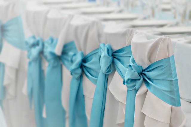 židle na svatbu s bílými potahy a modrými mašlemi, v pozadí bílý stůl